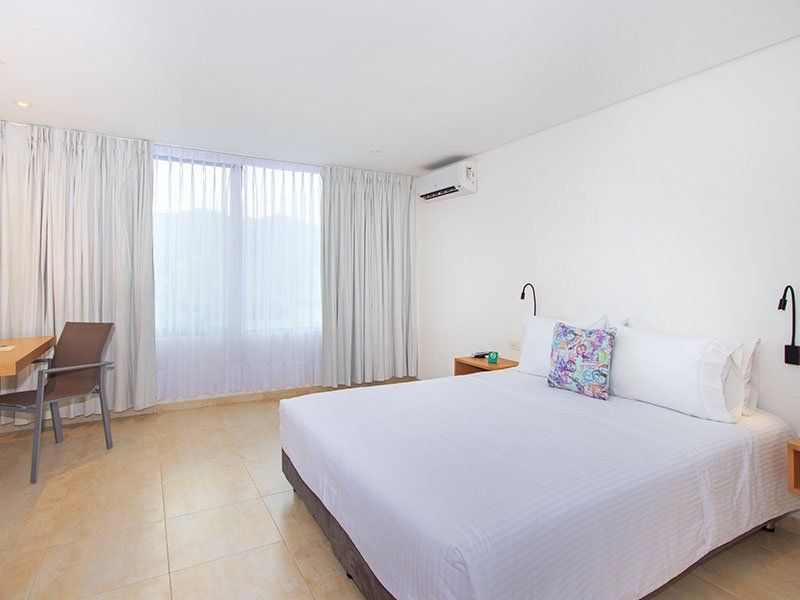 Habitación de hotel con cama, escritorio y ventana con cortinas blancas. Las paredes son blancas y el suelo es de baldosas.