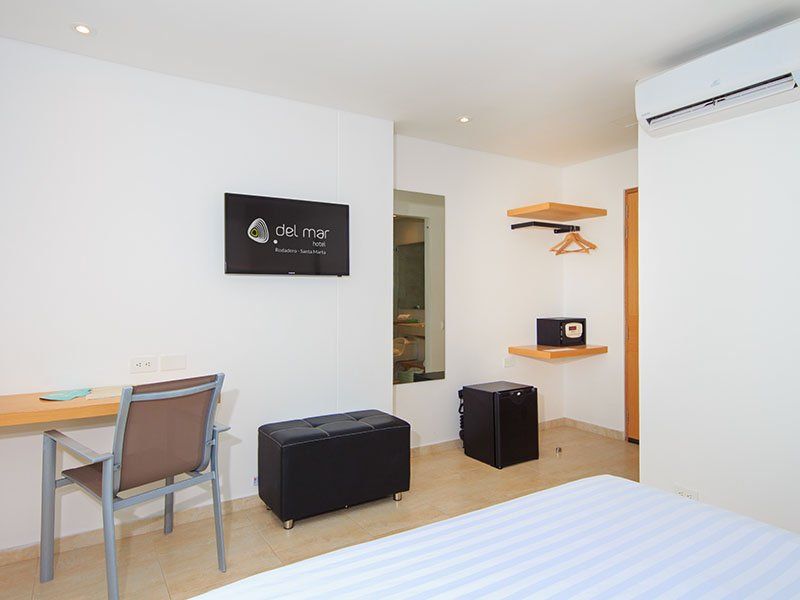 Habitación de hotel con cama, escritorio, TV, caja fuerte y mininevera. Paredes blancas y un diseño moderno y minimalista.