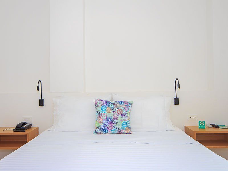 Un dormitorio de hotel minimalista con una cama blanca, una almohada colorida y dos mesitas de noche con lámparas.