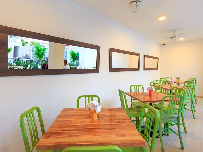 Un interior de restaurante bien iluminado, con sillas verdes y mesas de madera, espejos en paredes blancas y una vista de la vegetación reflejada en un espejo.