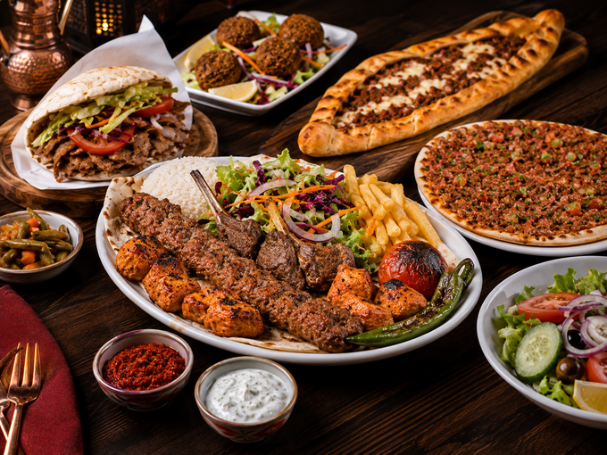 Un assortimento di piatti mediorientali con kebab, pane piatto, salse, insalata e foglie di vite ripiene, disposti su un tavolo.