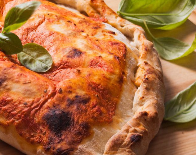 Primo piano di una pizza dorata con formaggio fuso e foglie di basilico su un tagliere di legno.