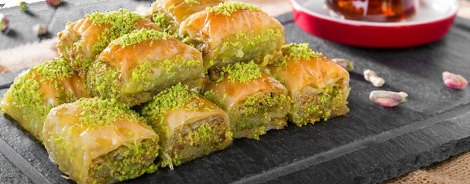 Vassoio di quadratini di baklava al pistacchio ricoperti di granella di pistacchio su un tagliere di ardesia scura, con sciroppo sullo sfondo.