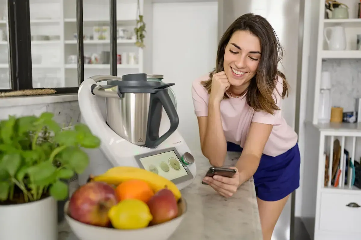 A woman is standing in front of a food processor and looking at her phone.