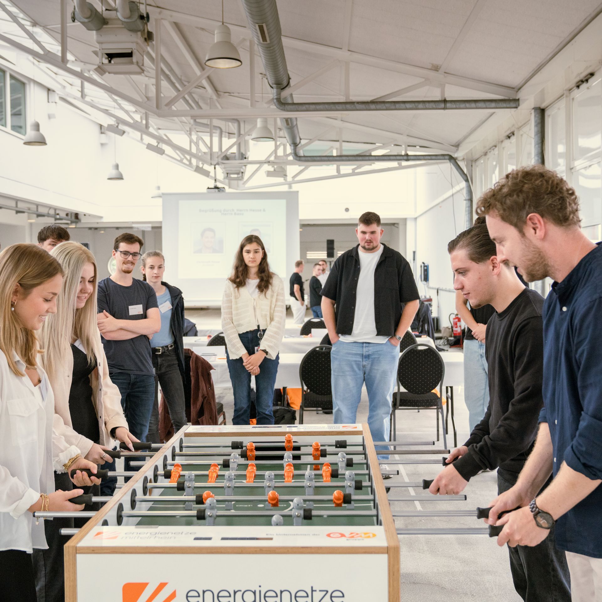 Eine Gruppe junger Frauen und Männer spielt in einem großen Raum Tischkicker. Am Kickertisch stehen links zwei Frauen, rechts zwei Männer, dahinter junge Menschen die zuschauen. 
