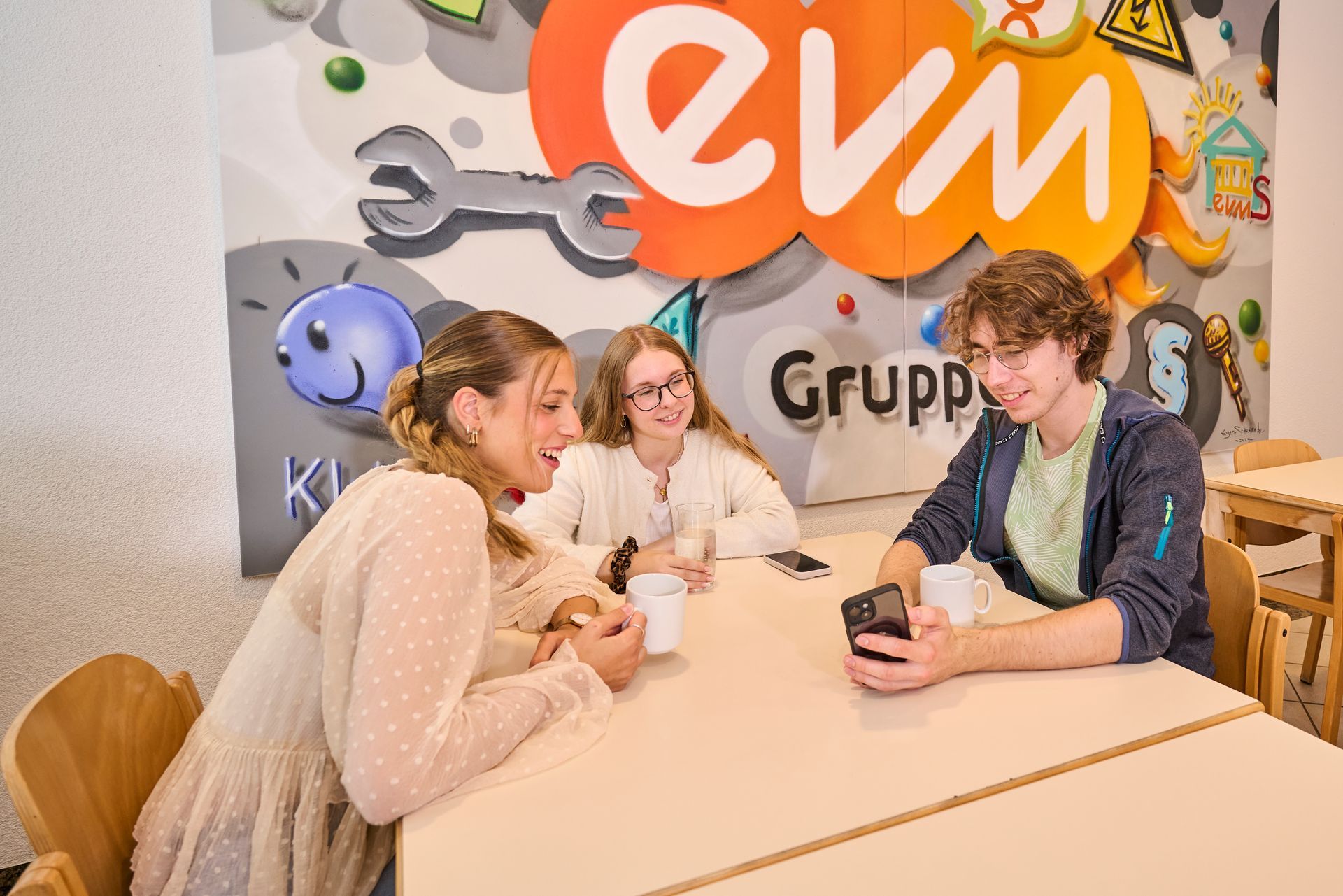 Drei junge Menschen sitzen mit Kaffe und Handy an einem Tisch, im Hintergrund evm Logo Wand. 
