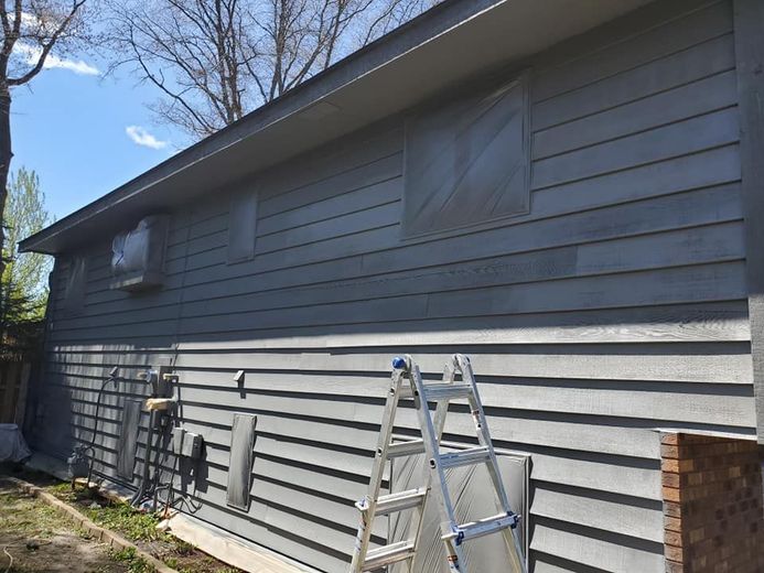 Grey-sided house with a ladder and covered windows during painting. Trees and blue sky visible.