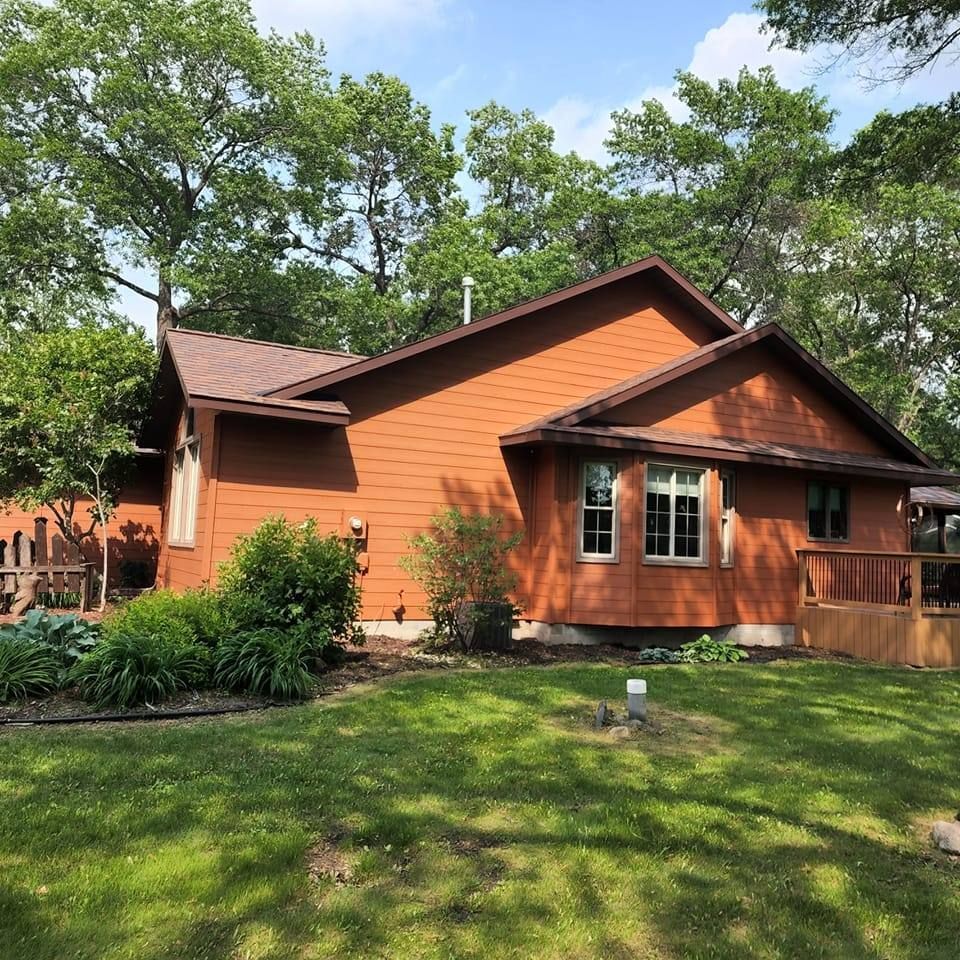 Brown house with a brown roof, green yard, and leafy trees on a sunny day.
