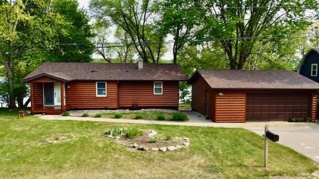 Brown ranch-style house with attached garage on a green lawn, set against trees and a body of water.