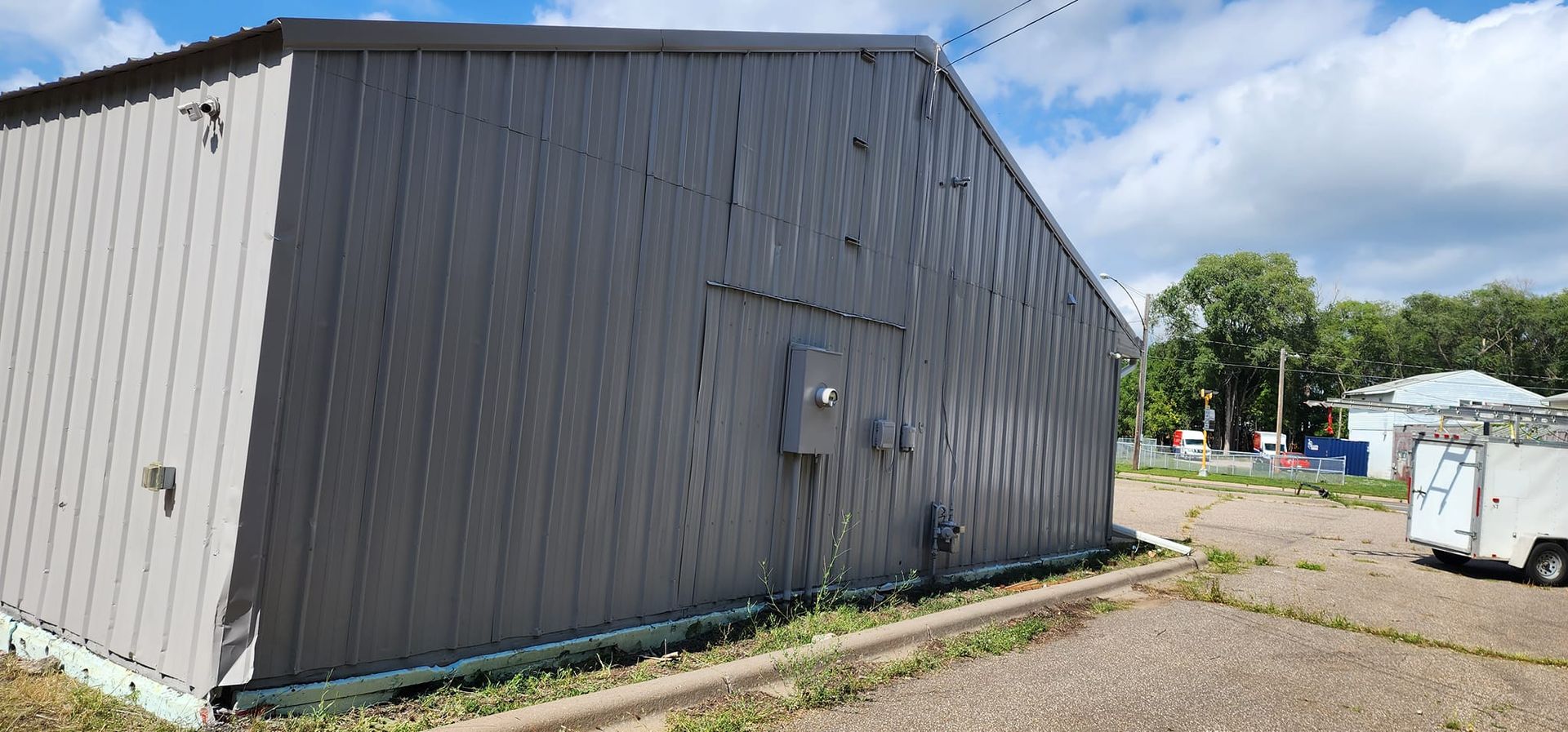 A gray metal building with a side view, near a gravel lot and trees under a cloudy sky.