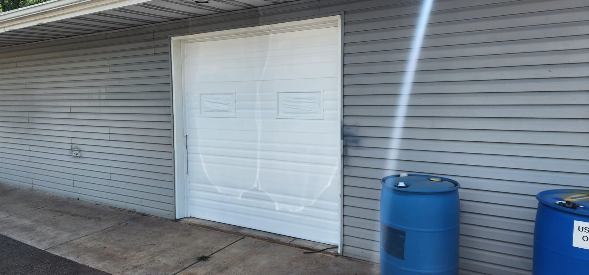 Exterior of a building with a large white door, blue barrels, and a beam of light.