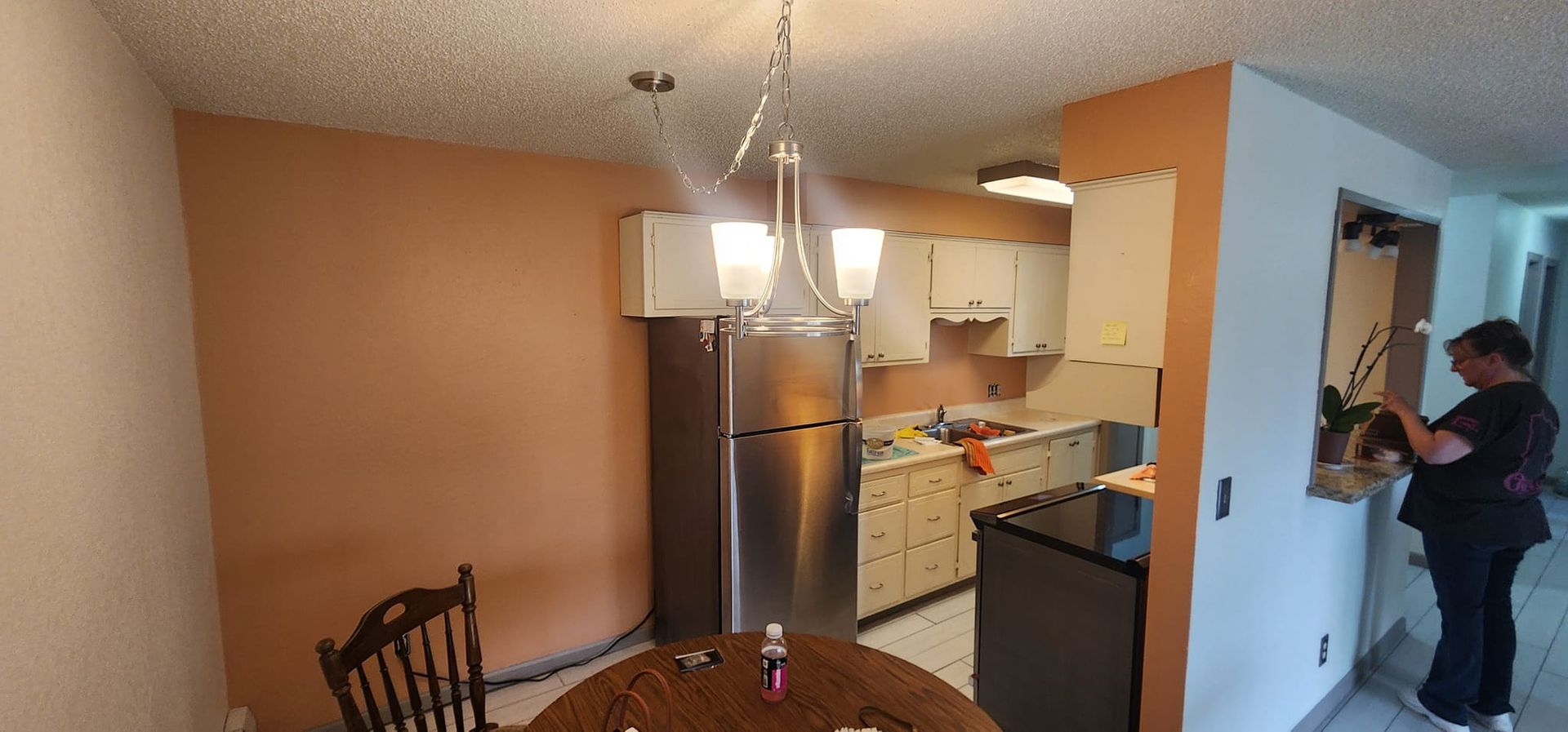A woman in black stands near a doorway in a home. The kitchen features a fridge and cabinets.