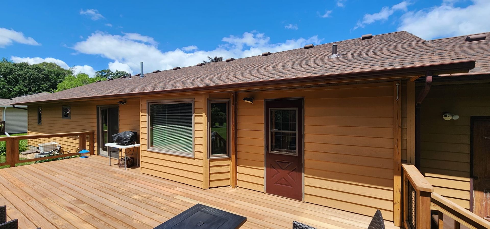 A house with a wooden deck under a blue sky with fluffy clouds. Brown roof, siding, and deck railing.