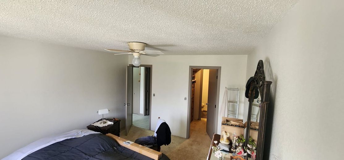 A bedroom with a bed, bathroom door, closet door, ceiling fan, and dresser. White walls and popcorn ceiling.