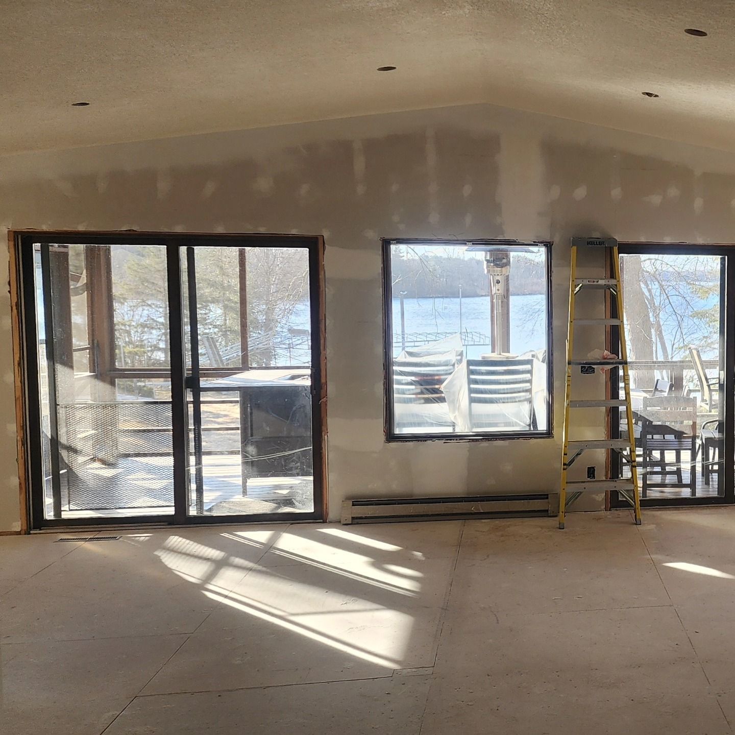 Interior of room under construction, with windows and a sliding glass door. Drywall unfinished, ladder present.