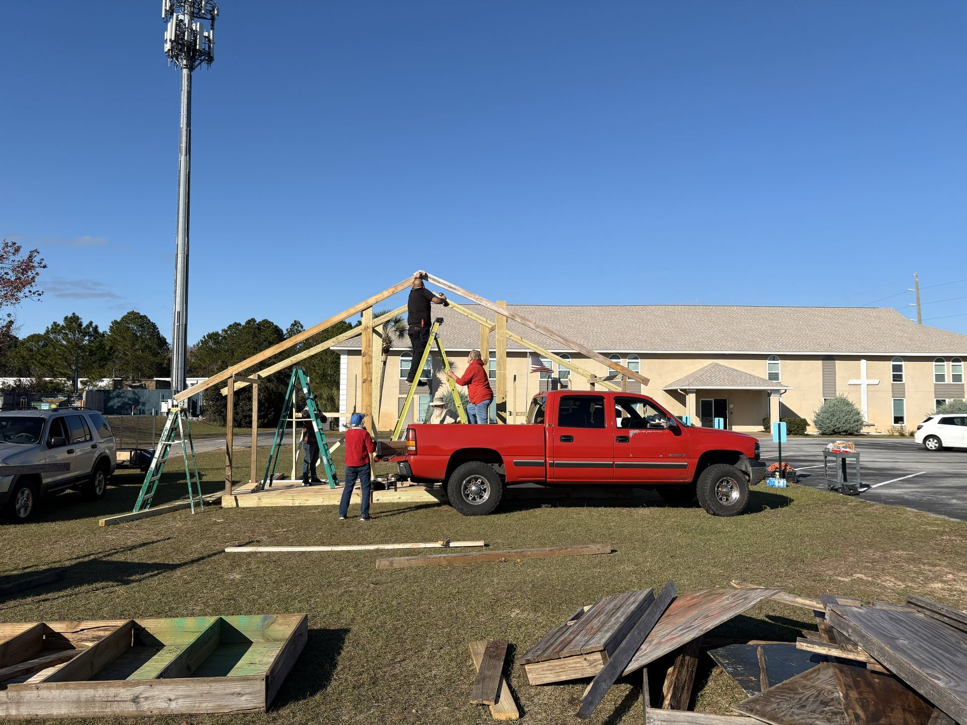 People constructing a wooden structure outside a beige building; red truck nearby.