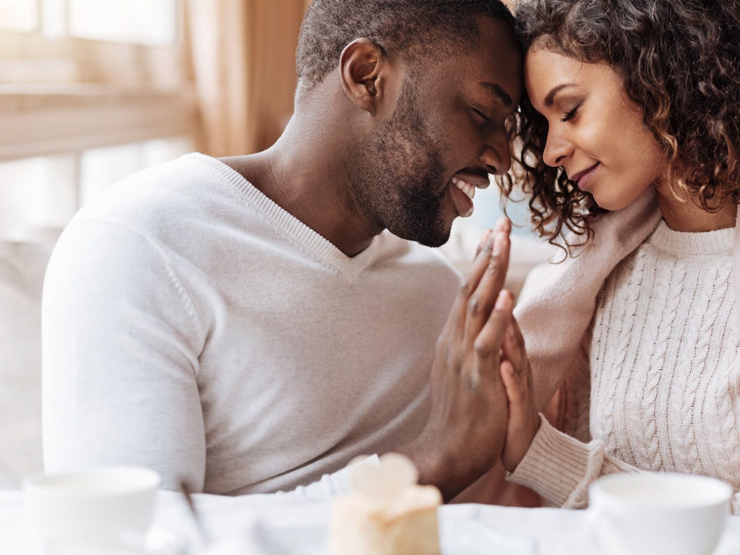 Man and woman in cozy setting, holding hands, foreheads touching, smiling.