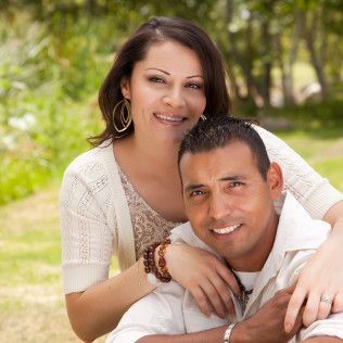 Couple smiling outdoors, woman's arm around man. They appear happy in a park setting.