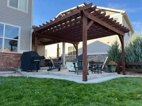 Brown pergola over a concrete patio with outdoor furniture, next to a house and green lawn.