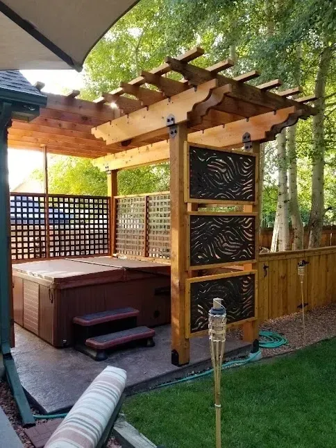 Hot tub under a wooden pergola with decorative privacy screens in a backyard setting.