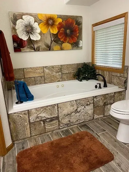 Bathroom with a built-in tub, stone surround, floral artwork, and a window with blinds.