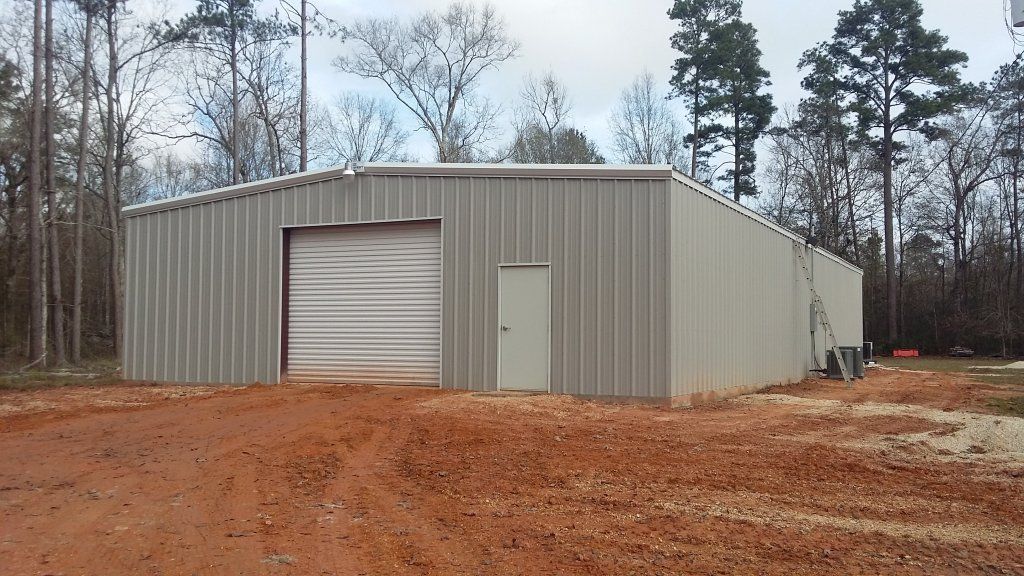 Large gray metal storage building with roll-up door on a dirt lot, surrounded by trees
