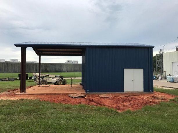 Blue metal shed with open shelter, white door, and equipment on red dirt in a grassy field