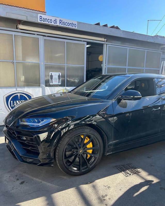 Un SUV Lamborghini Urus nero con pinze dei freni gialle parcheggiato fuori da un edificio con la scritta 