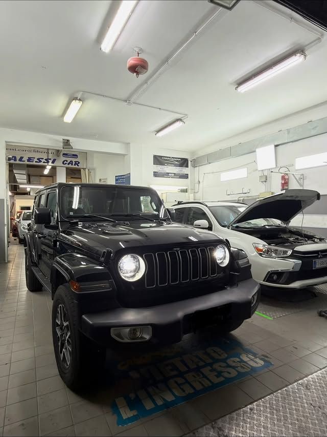 Una Jeep Wrangler nera in un garage, accanto a un'auto bianca con il cofano aperto. Luci fluorescenti in alto.