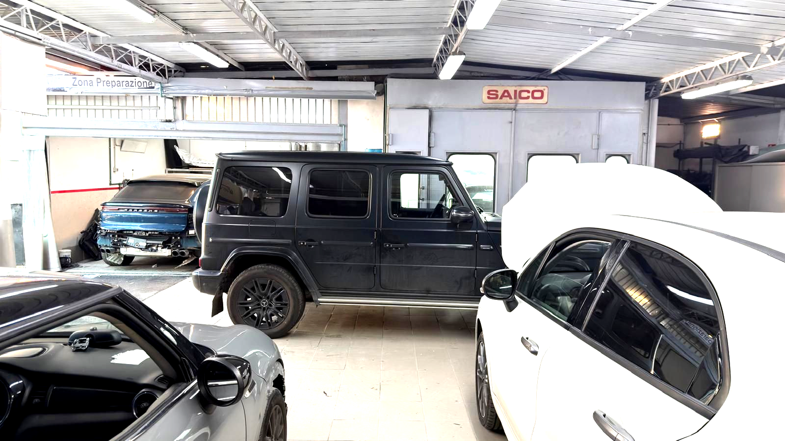 Auto bianca in carrozzeria con le portiere aperte, operaio vicino a un'altra auto nella cabina di verniciatura.