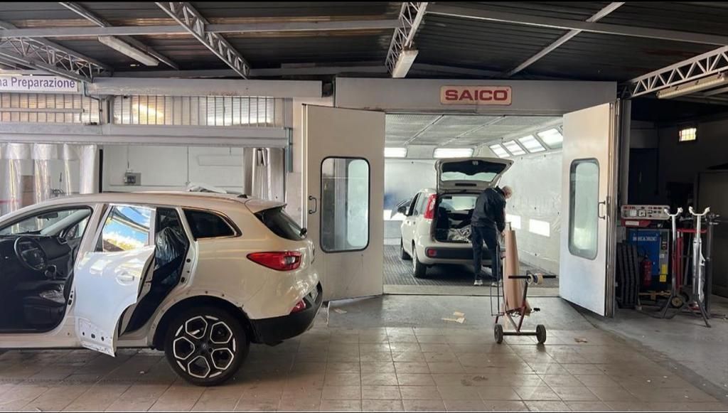 Auto bianca in carrozzeria con le portiere aperte, operaio vicino a un'altra auto nella cabina di verniciatura.