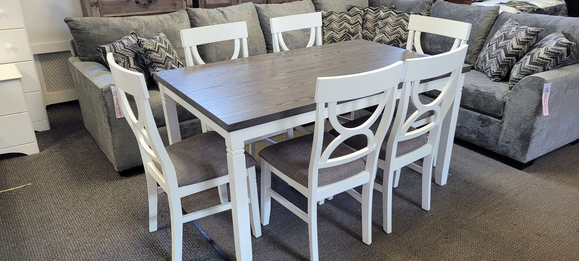 Dining table set with six chairs in front of a gray sectional sofa in a store.