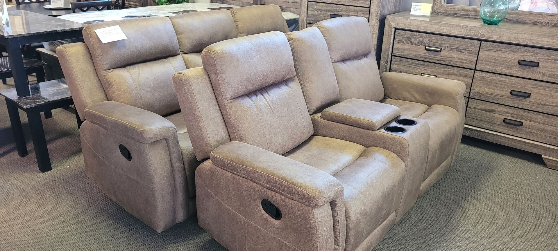 A two-seat tan recliner with a center console and cupholders sits in a furniture store.