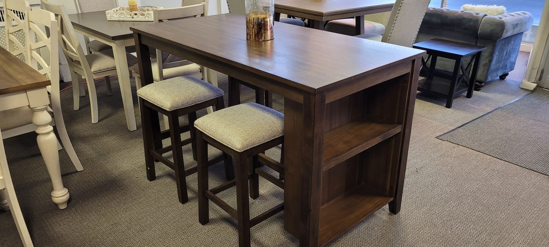 A dark wood bar table with two stools. It has a shelf on the end and a glass on top.