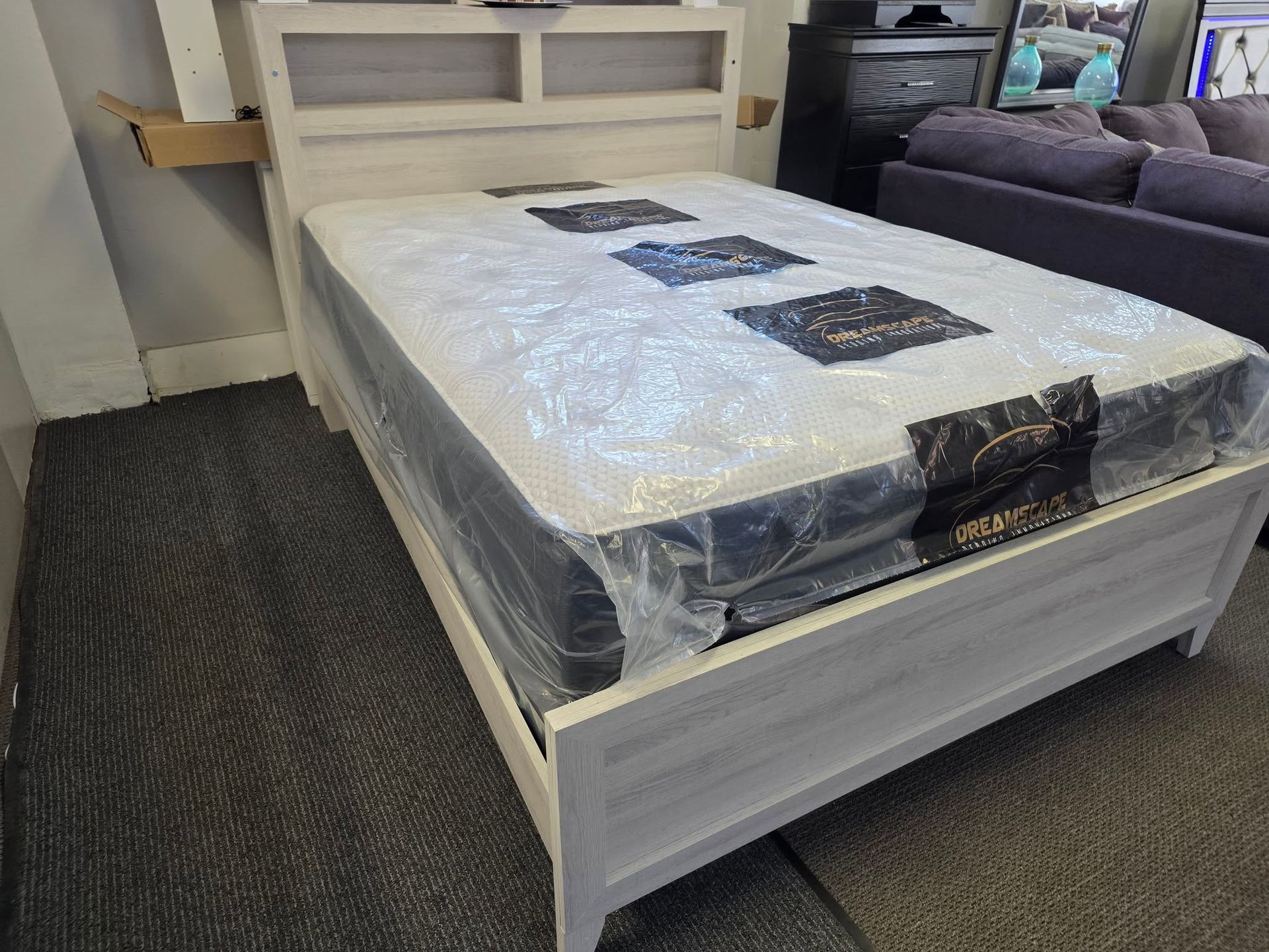 A white bed with a headboard, mattress covered in plastic, in a showroom.