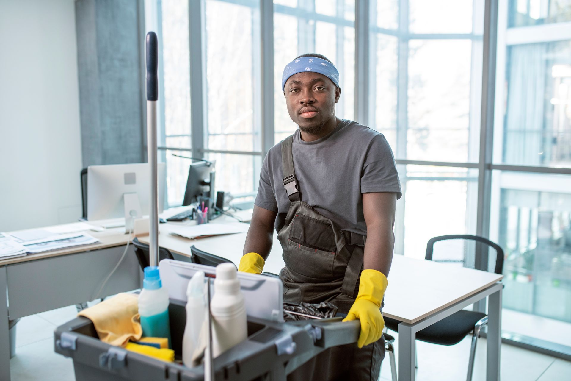 A man is sitting at a desk in an office holding a mop and cleaning supplies.
