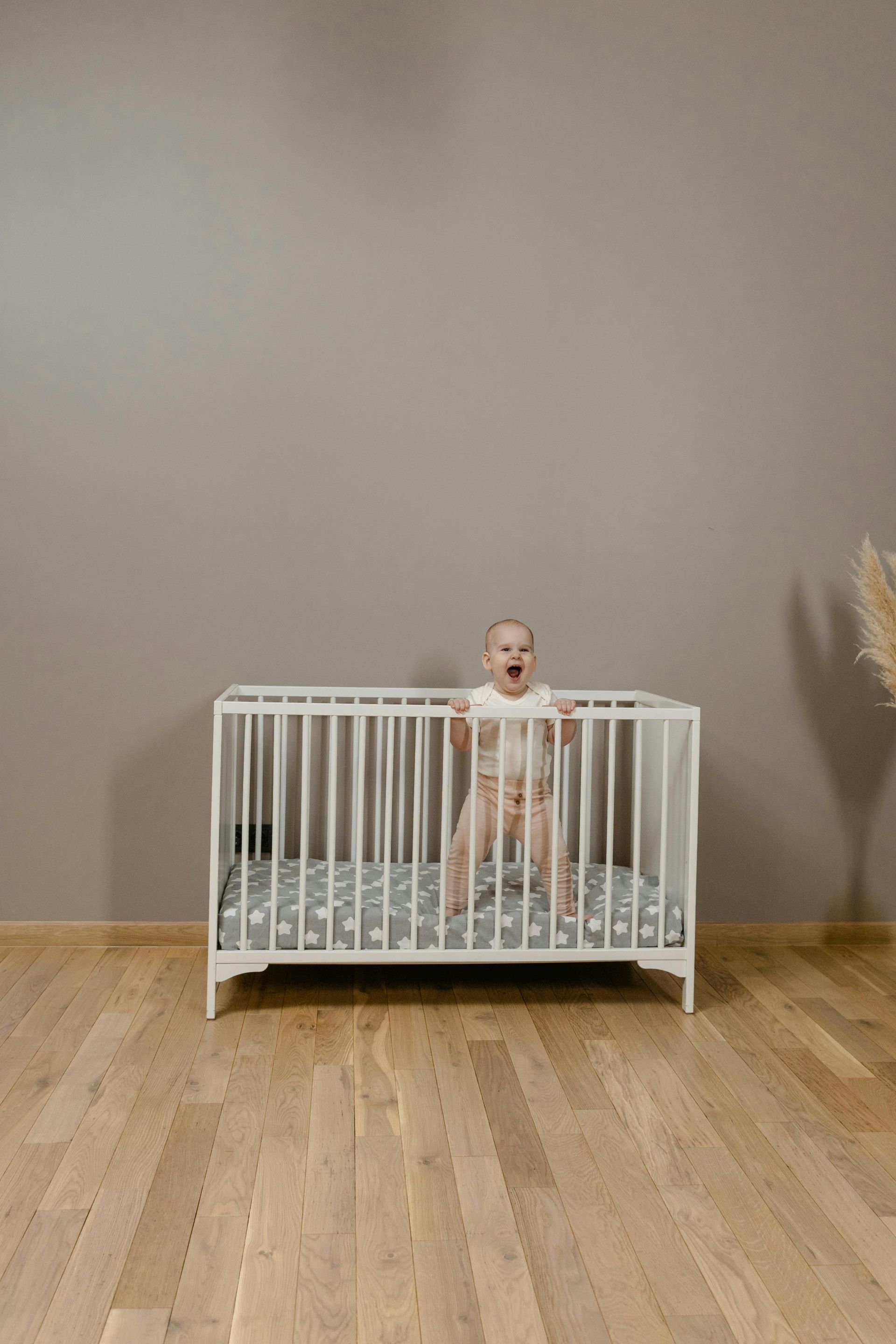 A baby is standing in a white crib on a wooden floor