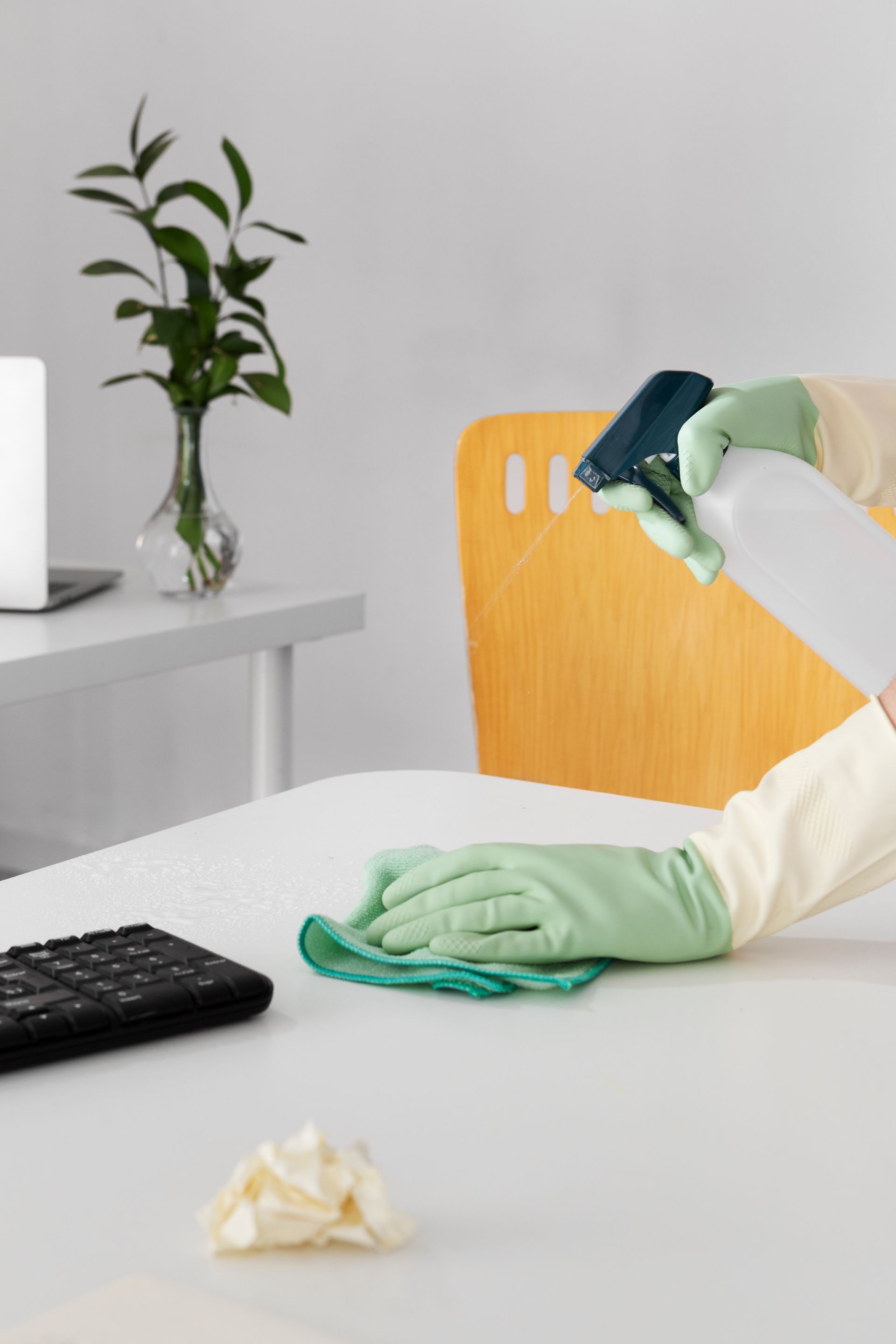 A person wearing green gloves is cleaning a desk with a spray bottle
