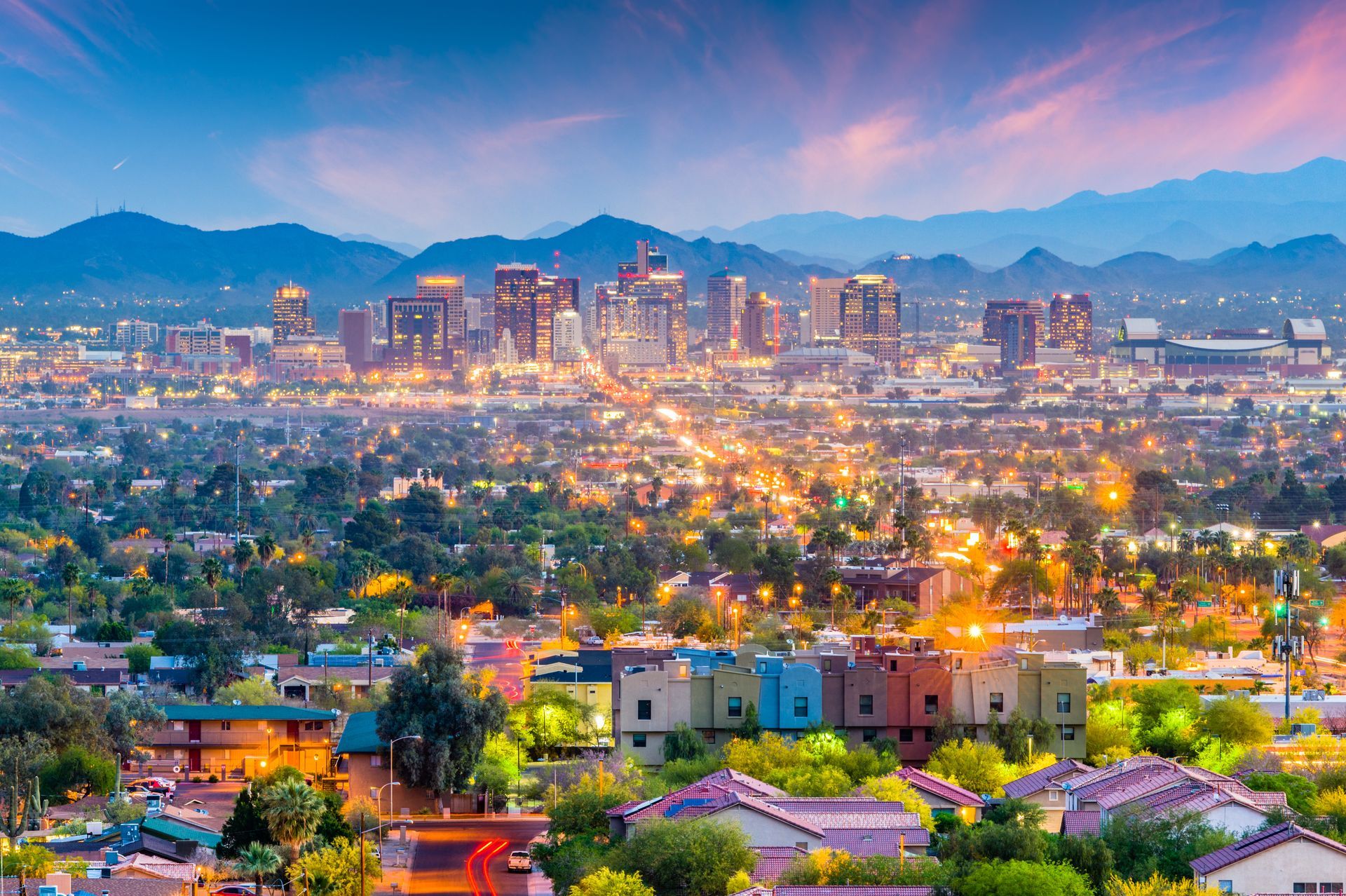 City skyline at dusk with colorful lights, mountains in the distance, and houses in the foreground