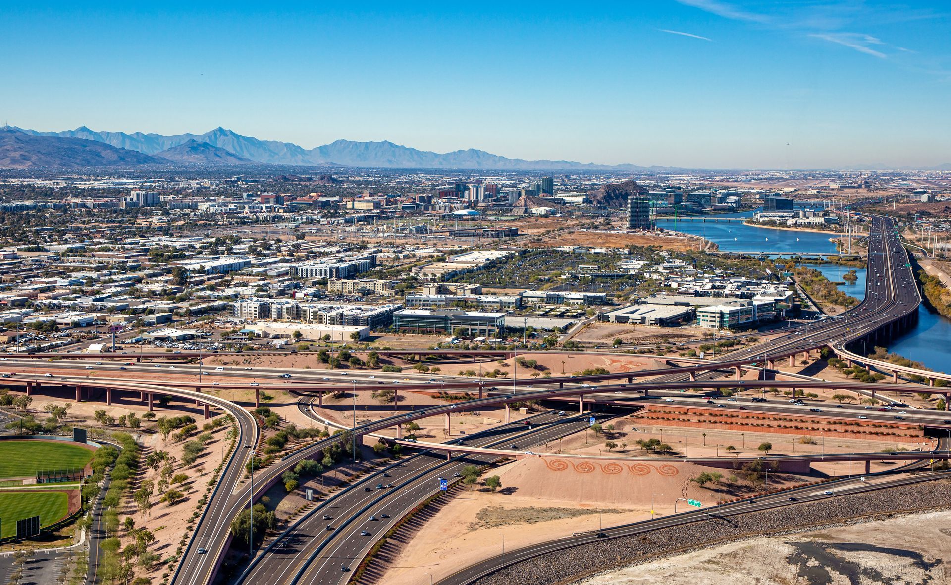 Aerial view of a cityscape with highways, buildings, and mountains under a clear blue sky.