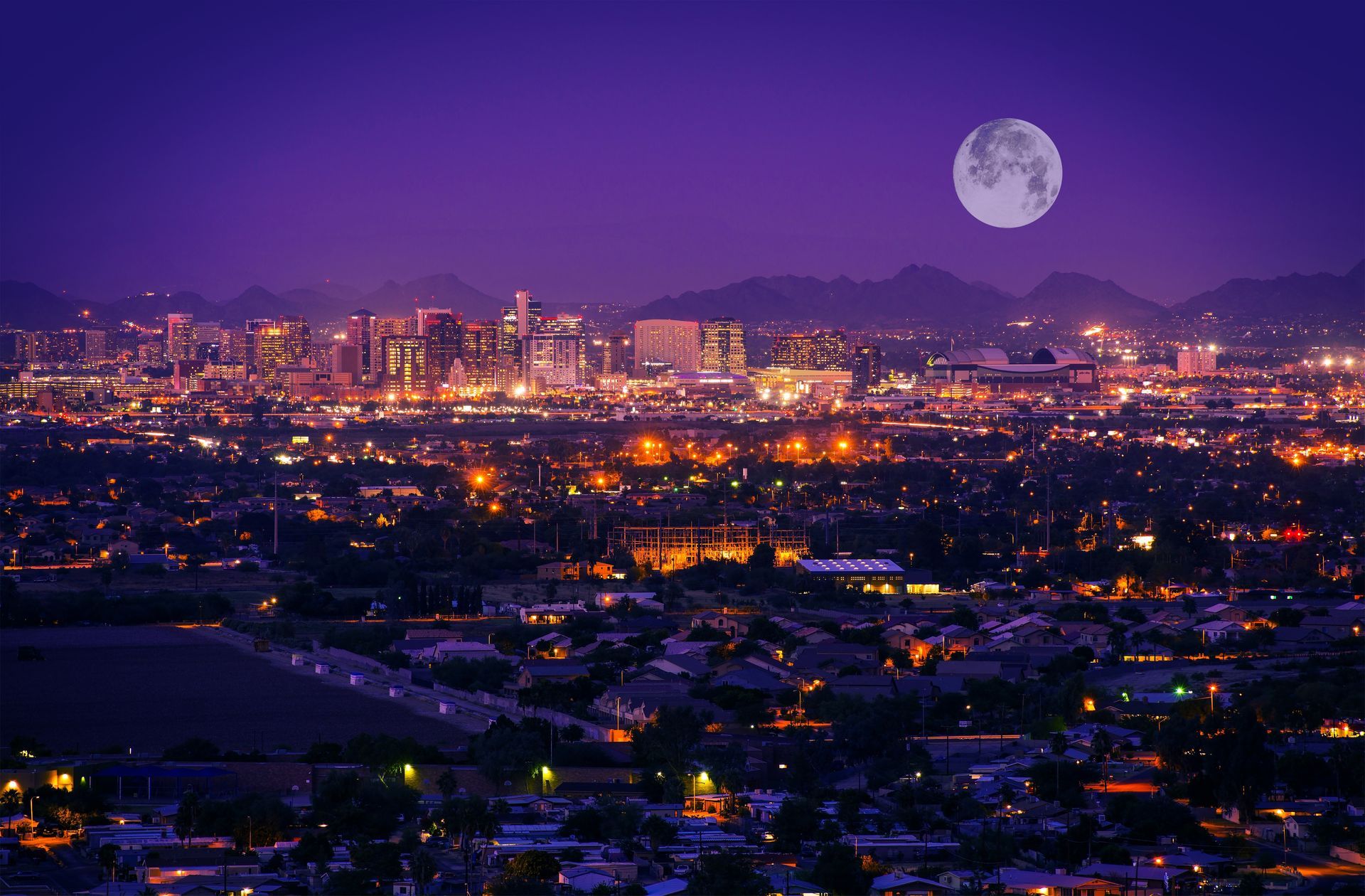 Moonlit city skyline at dusk with glowing lights and purple mountains in the background