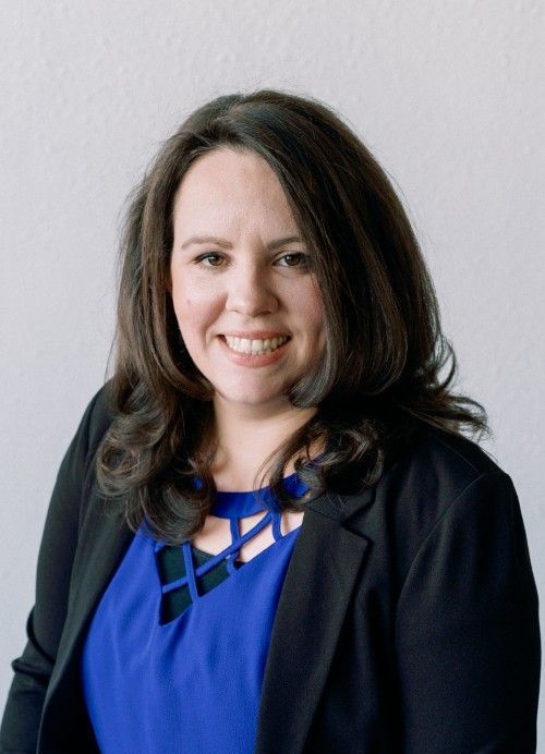 A woman in a blue dress and black jacket is smiling for the camera.