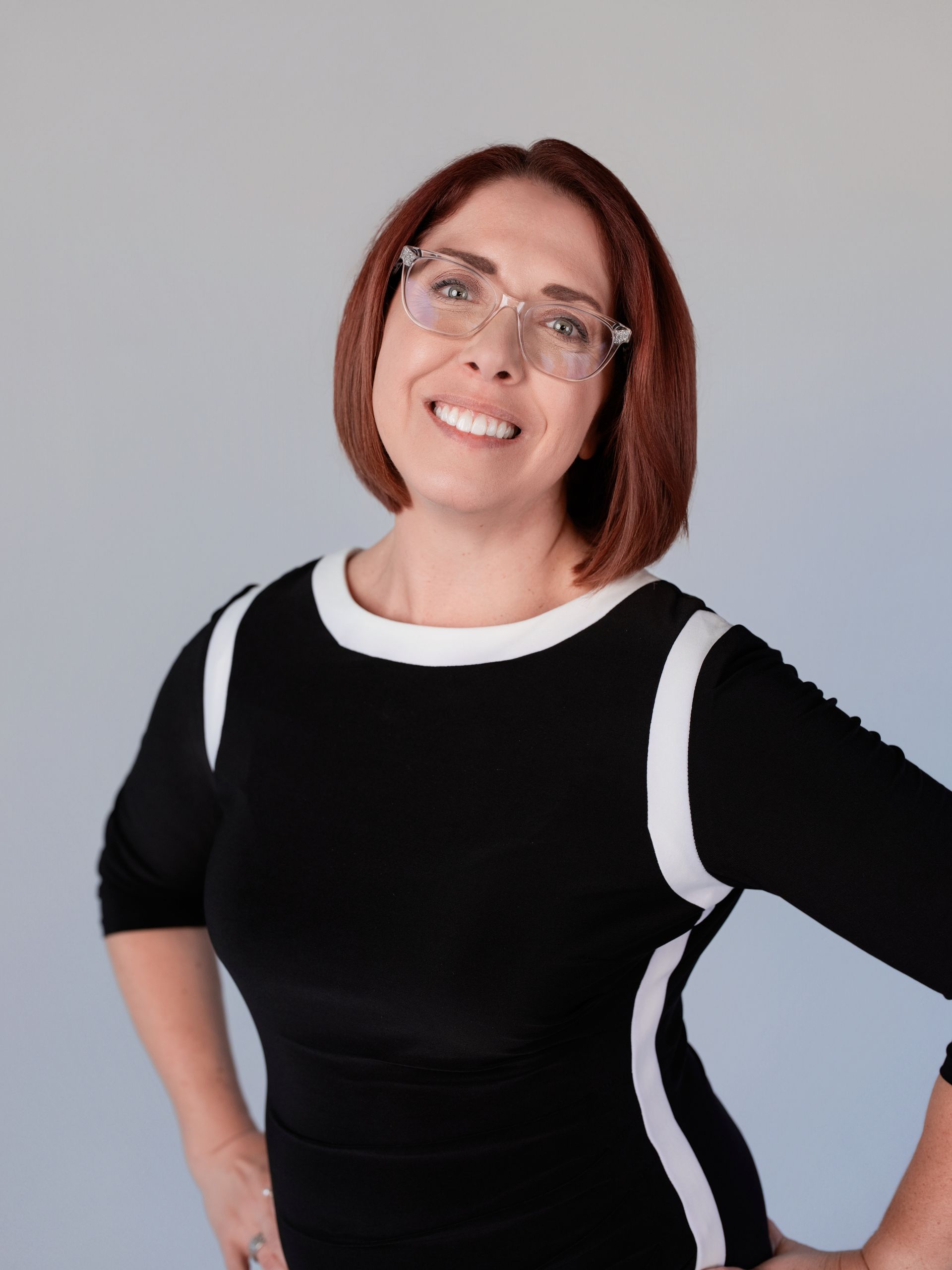 Woman with red hair, wearing glasses, and a black dress with white trim, smiling.
