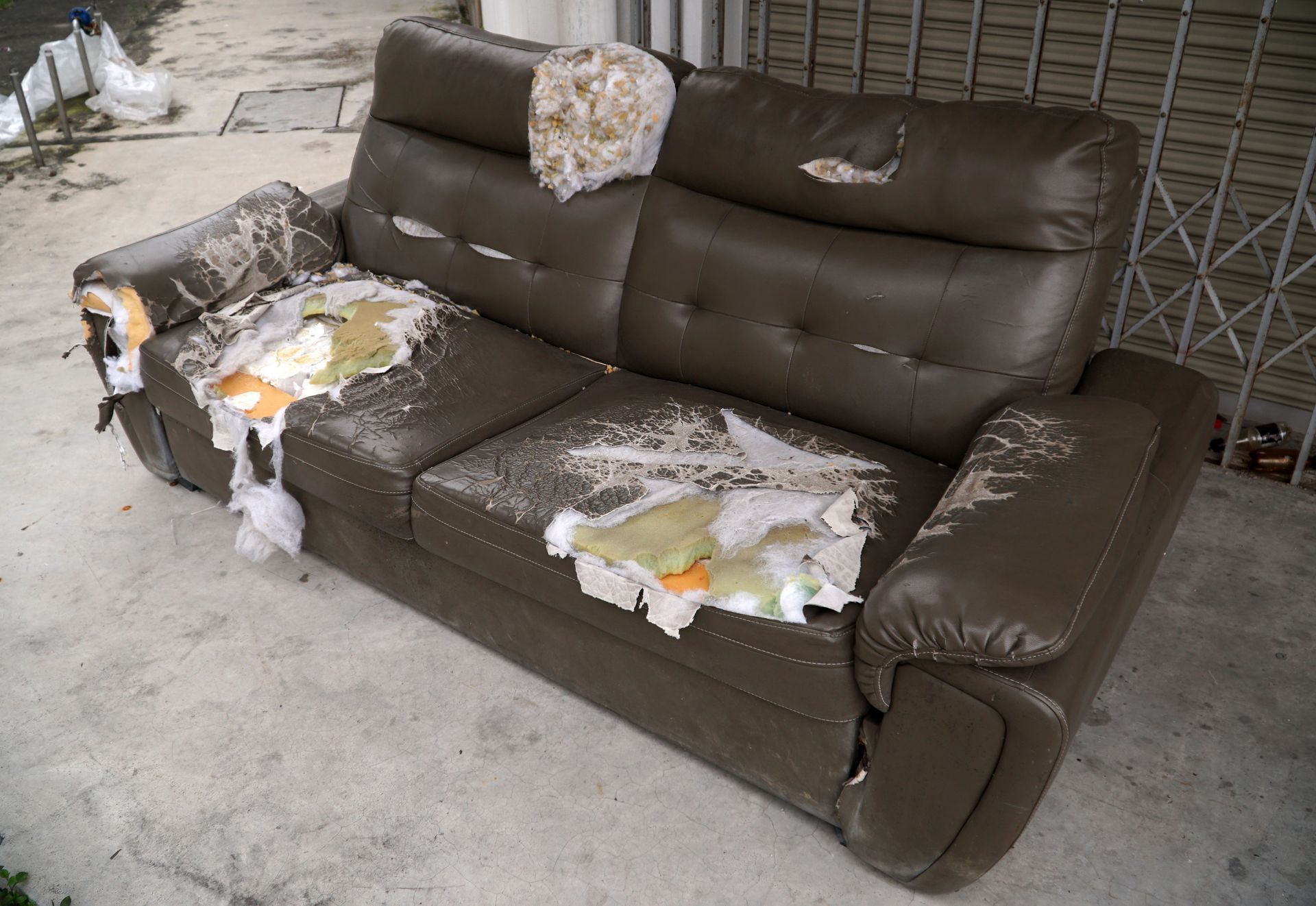 Damaged brown leather sofa with tears exposing stuffing, sitting on concrete.