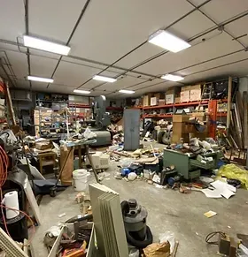 Messy workshop filled with equipment, boxes, and debris; overhead fluorescent lights.
