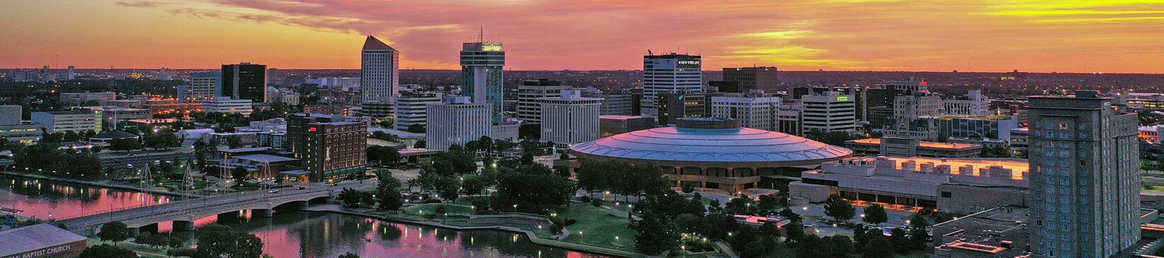 City skyline at sunset with colorful sky. Buildings, water, and bridge are visible.