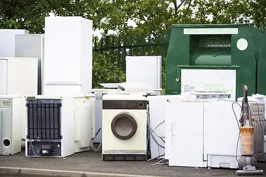 Discarded appliances, including a washing machine, refrigerators, and a vacuum cleaner, near a recycling bin.