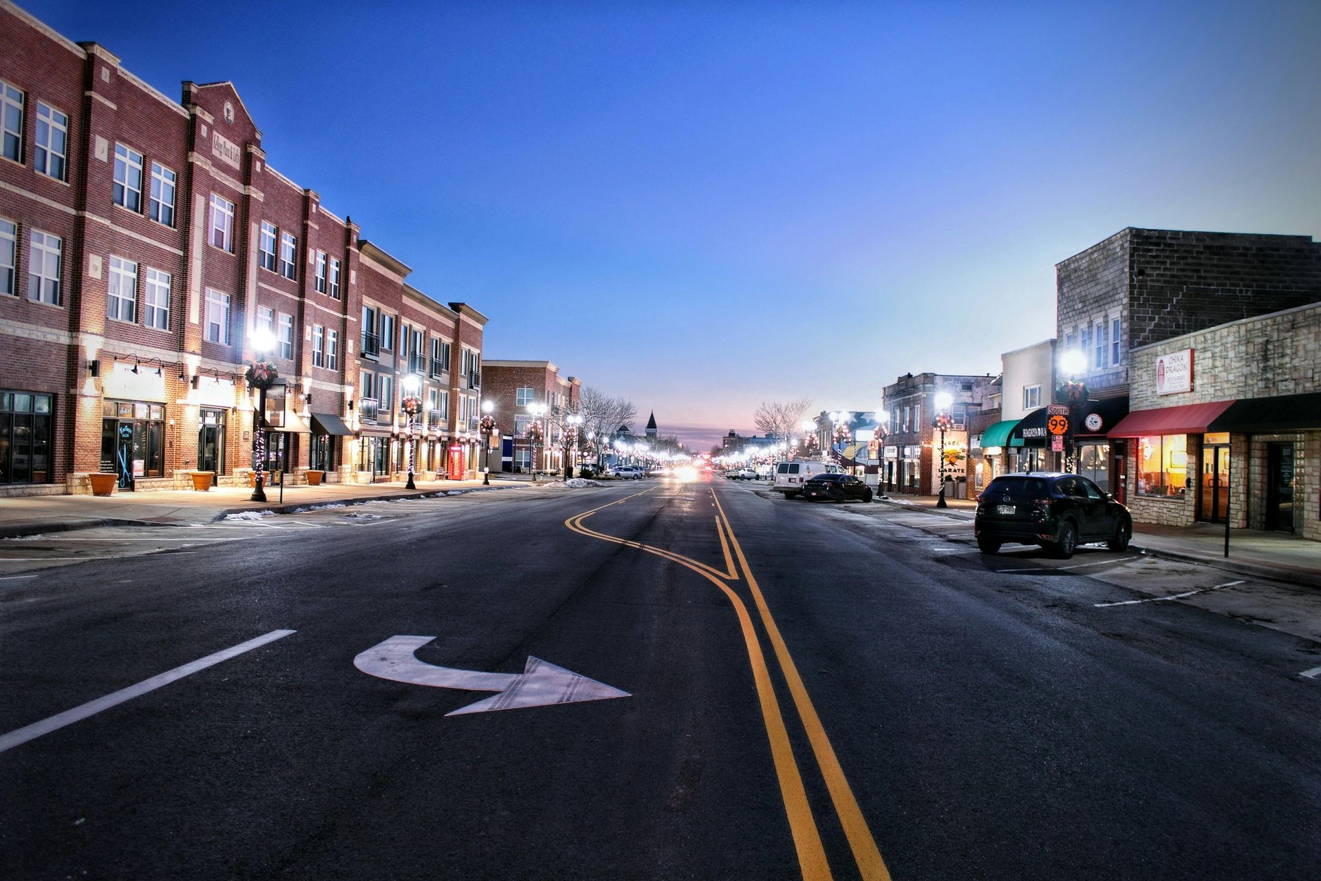 Street view at dusk, buildings with lights line a curved road. An arrow indicates the turn.