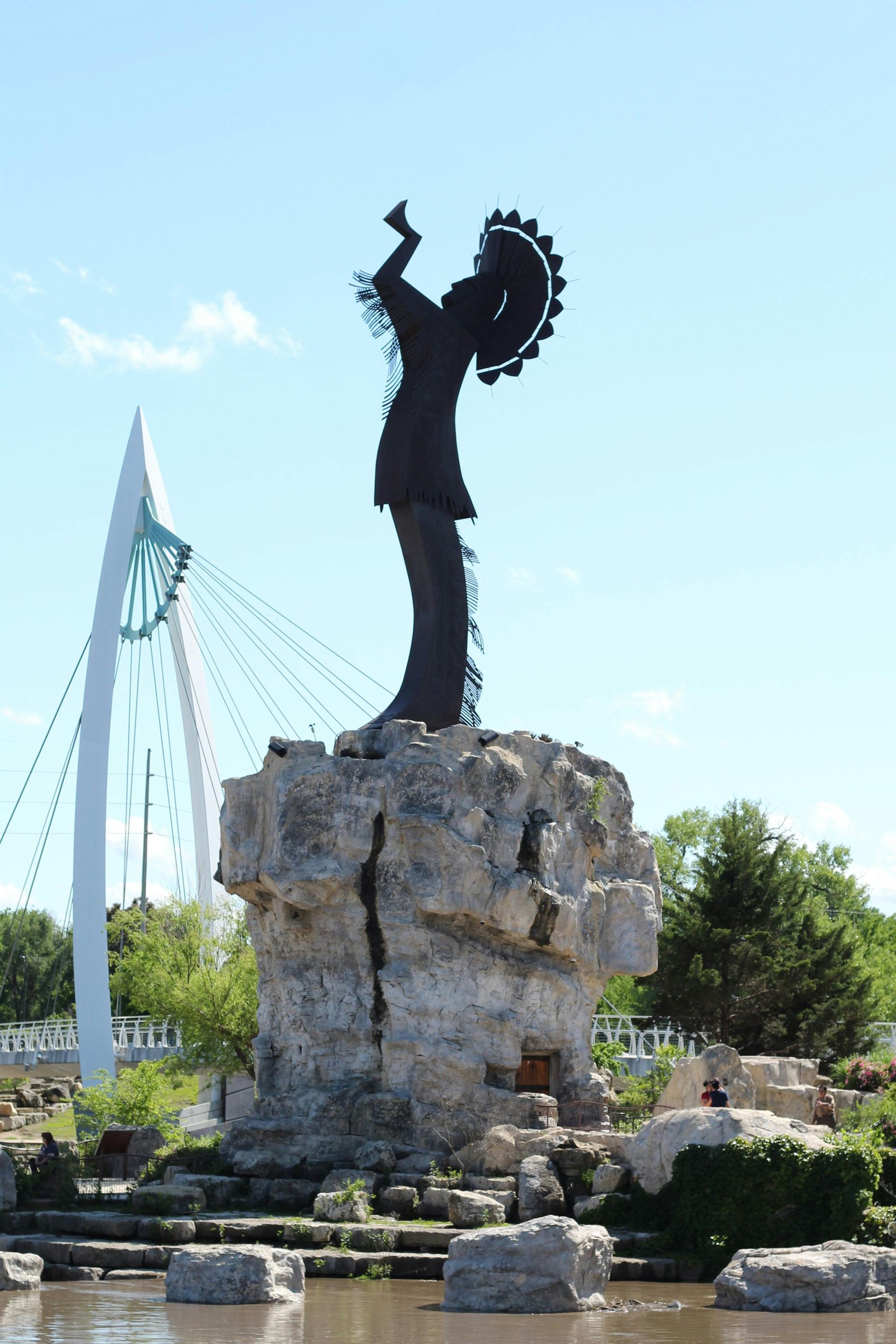 Sculpture of an Indigenous person with a headdress on a rocky base, in front of a modern bridge.