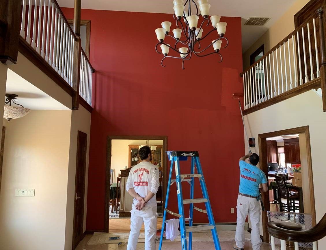 Workers painting a tall red wall in a foyer, with a blue ladder and chandelier overhead
